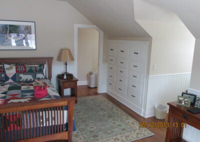 A cozy bedroom featuring a wooden bed with a quilt, a bedside table with a lamp, a patterned rug, a built-in wardrobe, and a wall picture.