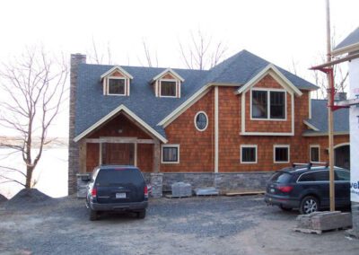Newly constructed house with cedar shingle siding and stone foundation, with two parked suvs in the unfinished driveway.